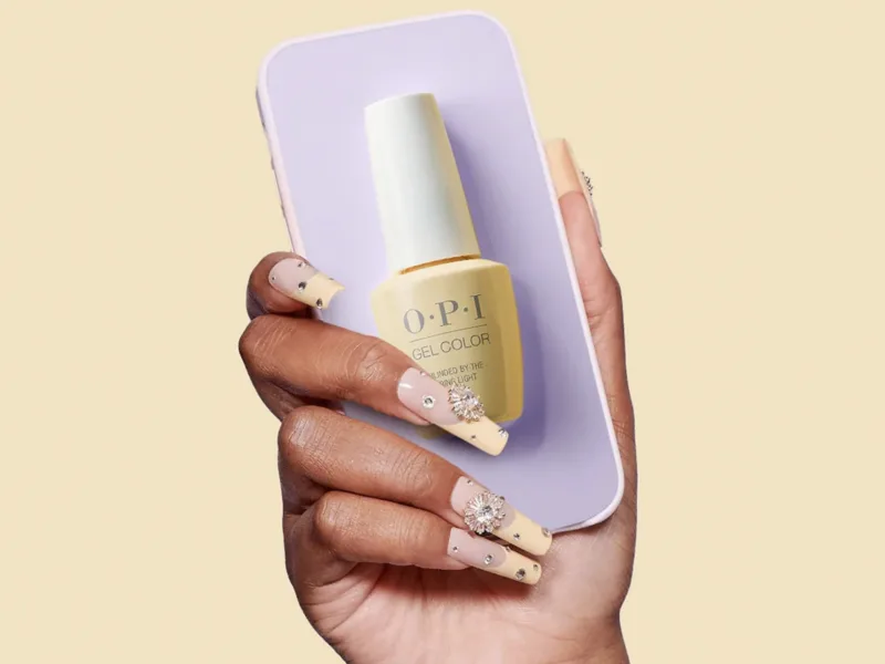 A woman with long yellow bejeweled nails holding OPI nail polish highlighting shades of yellow and white.