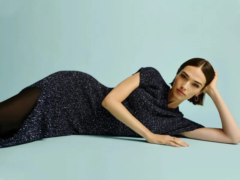 A woman in a sleek sparkly black dress reclines gracefully on the floor, against a turquoise backdrop.