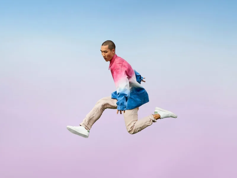 A man in mid-air jumps against a pastel gradient sky background, wearing a tie-dye dress shirt and khaki pants.